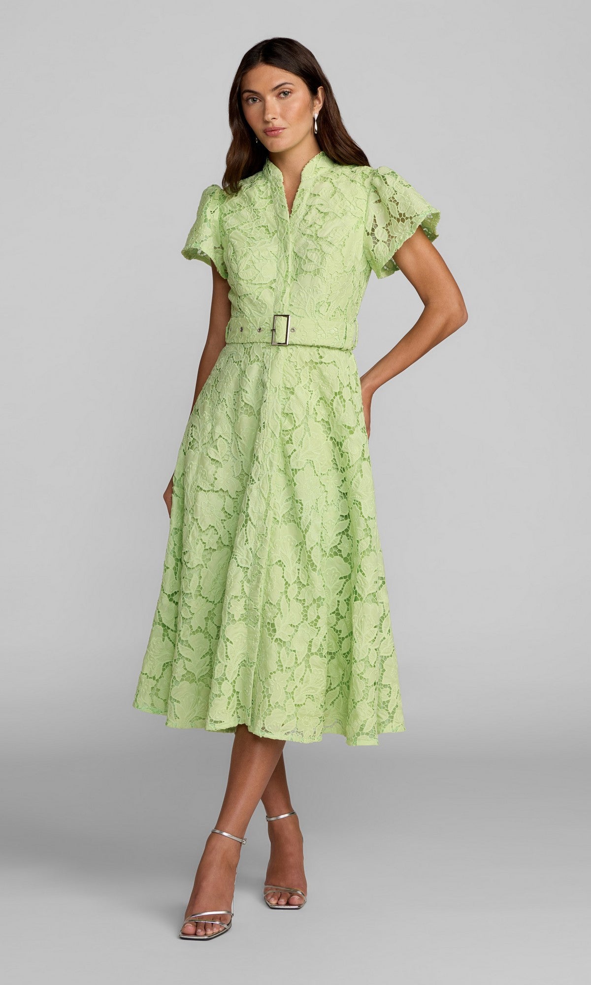 A woman stands in the Mac Duggal 56320 Midi Party Dress—a sage lace-embroidered dress with short sleeves and a matching belt—paired with silver high-heeled sandals.